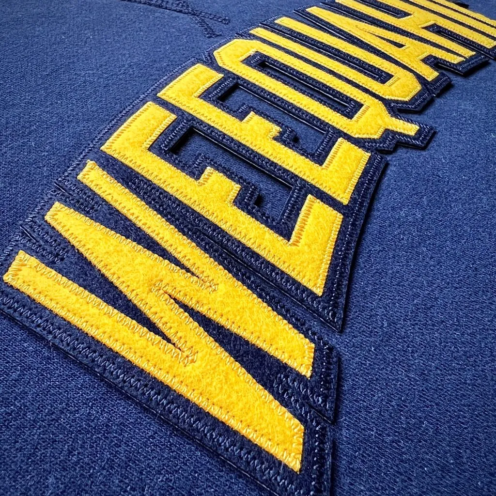 Gold tackle twill letters with a navy satin stitched border layered onto a navy athletic jersey