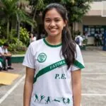 OLFU PE uniform t-shirt: white shirt with Fatima University seal on the chest, FATIMA diagonal sash across the torso, and P.E. silhouettes on the lower hem